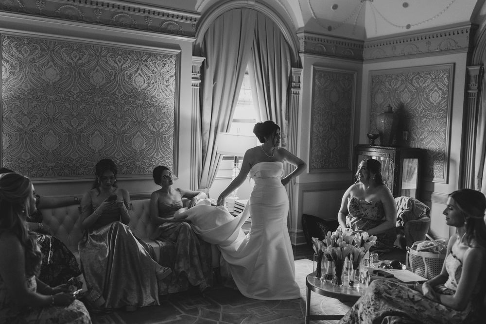 Wedding party enjoys candid black and white moment in an elegant room with ornate wallpaper as figure stands in flowing gown.
