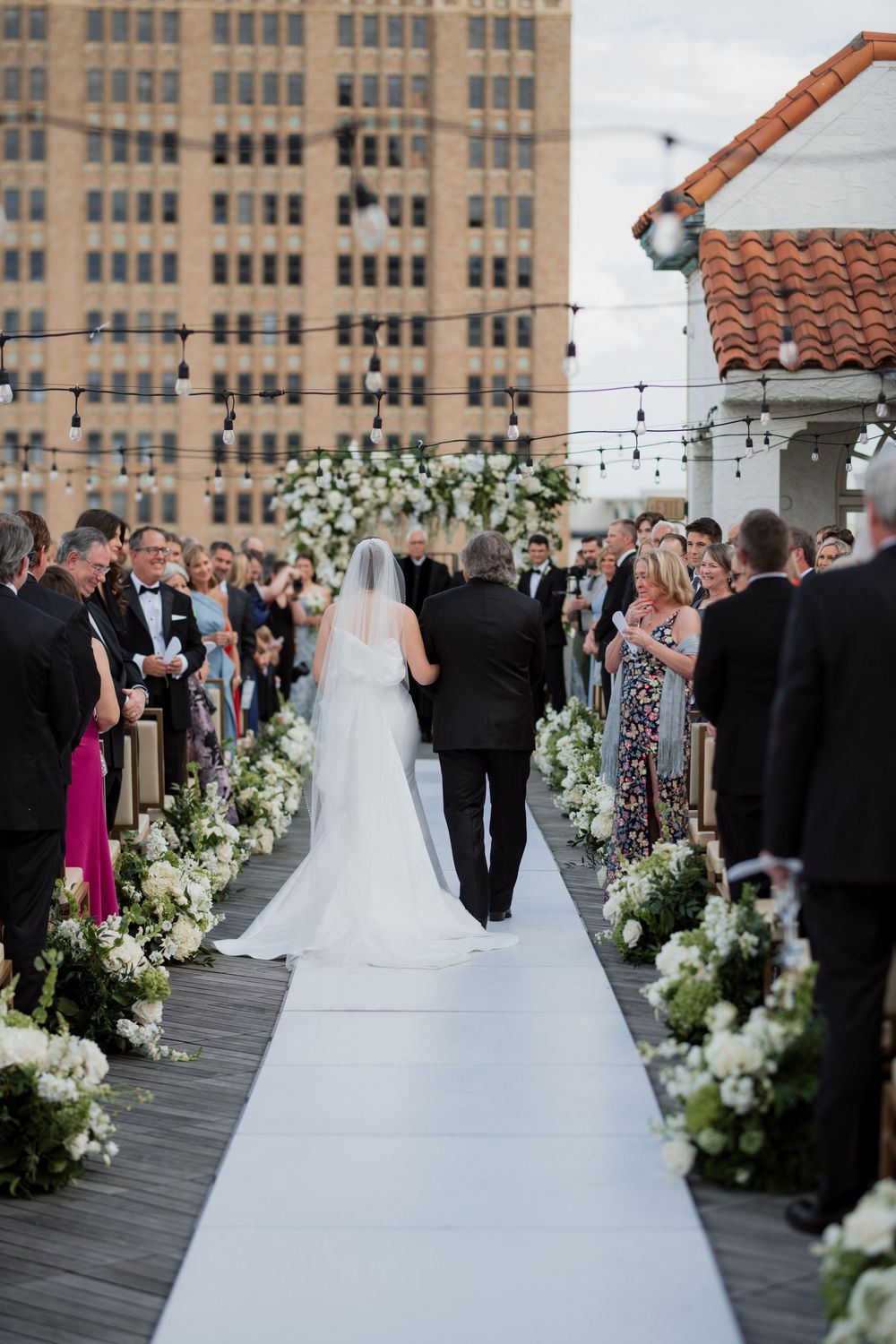 An elegant rooftop wedding ceremony with a white aisle runner bordered by lush floral arrangements against a city backdrop.