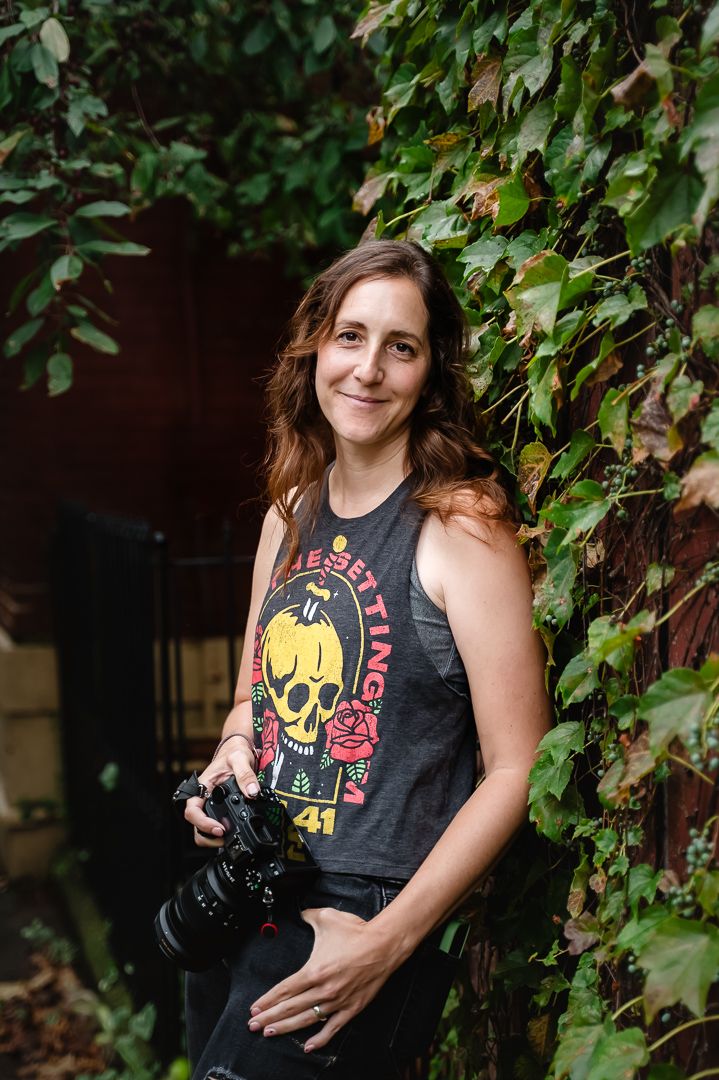 Female birth photographer smiling while holding Nikon camera outdoors.