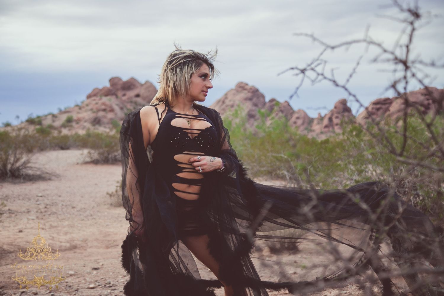 Woman in black shredded dress and flowing cape poses in desert landscape with red rock formations.