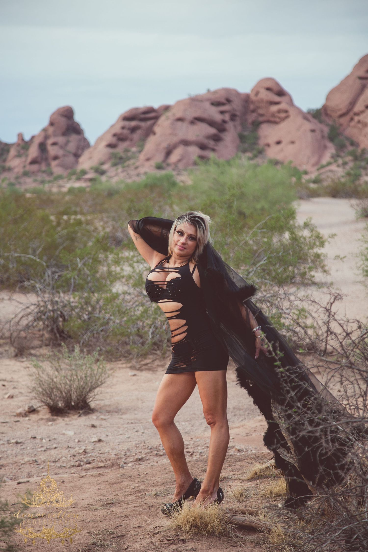 Woman in black dress poses by desert rocks with red sandstone formations and shrubs in background.