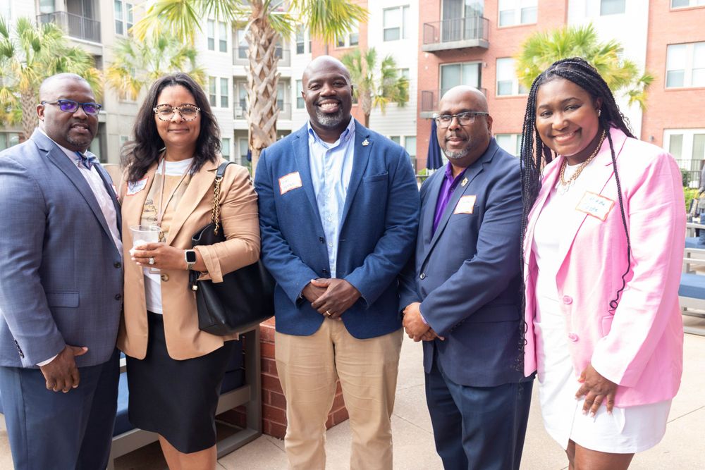 A group of professionals in business attire pose together outdoors with palm trees in the background.