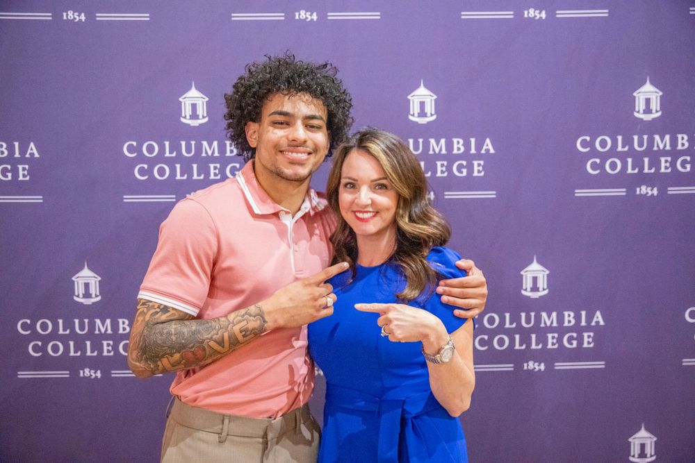 Two people posing in front of a purple branded backdrop with Columbia College logos.