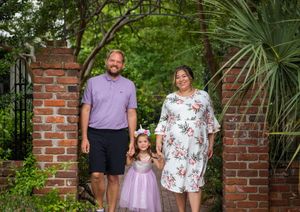 Three people pose for a family photo in a lush garden setting framed by brick pillars and tropical plants.