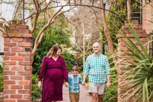 A family walks together through a garden path framed by brick pillars and lush greenery on a sunny day.