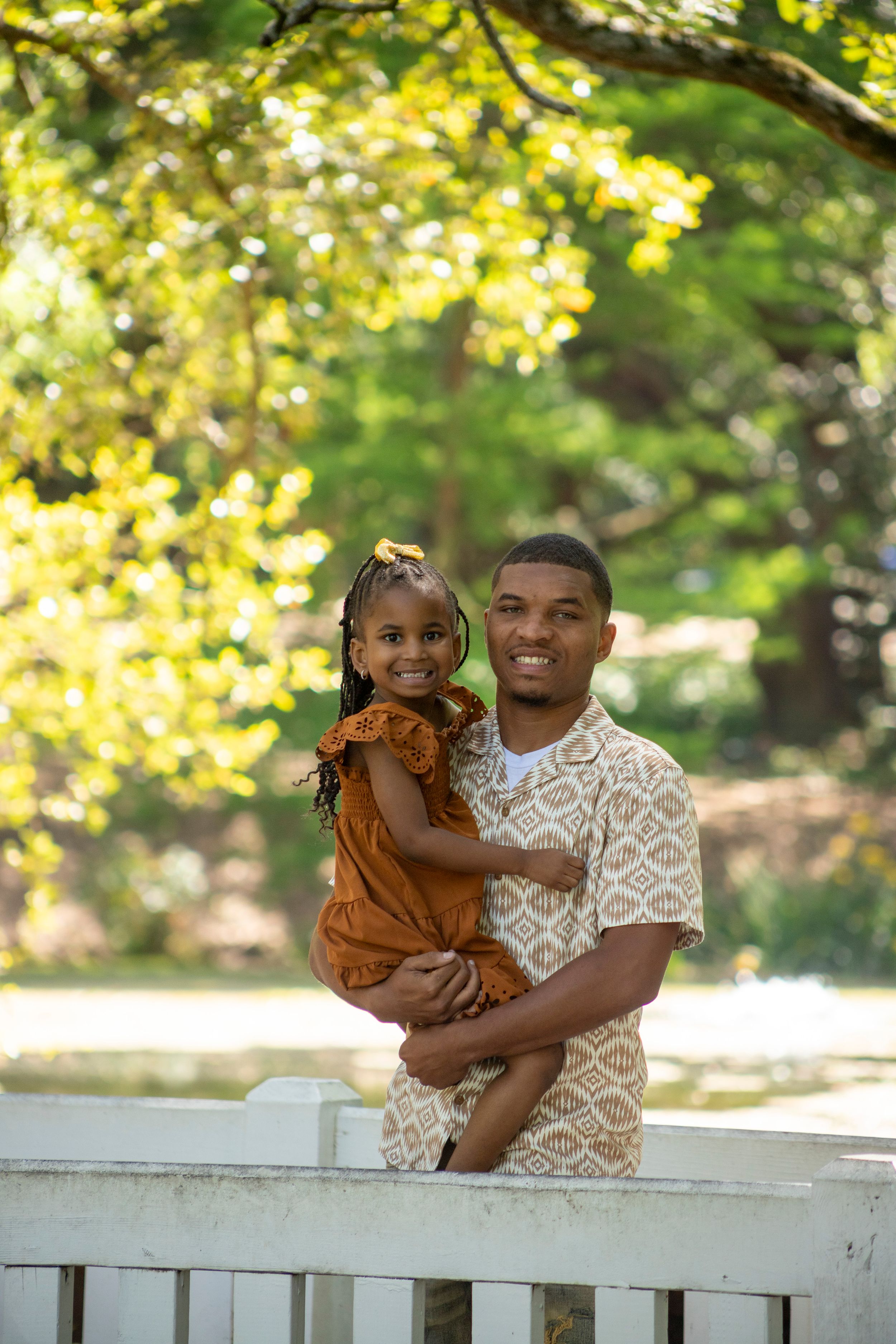A father and daughter enjoying a sweet moment together on a white railing with golden sunlit trees in the background.