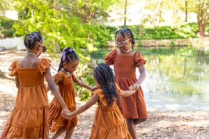 Four girls in matching rust-colored ruffle dresses walk together near a serene pond on a sunny day.