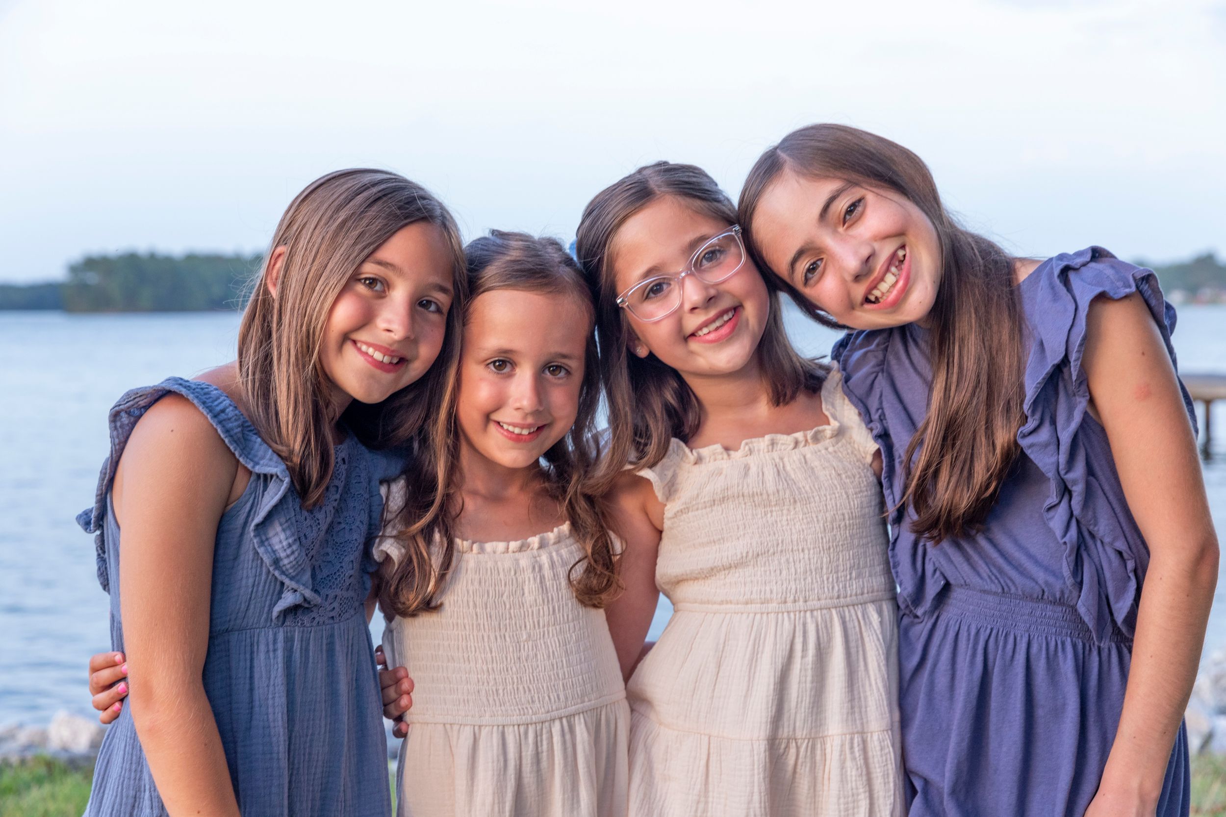 Four young girls in summer dresses smiling together by the lake on a sunny day.