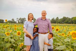 A family stands together in a bright yellow field of blooming sunflowers on a summer evening.