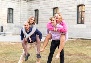 Two couples giving piggyback rides outdoors while smiling and having fun in front of a stone building.