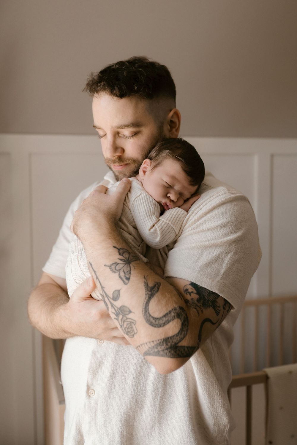 A tattooed parent in white holds a sleeping newborn against their chest in a minimalist room.