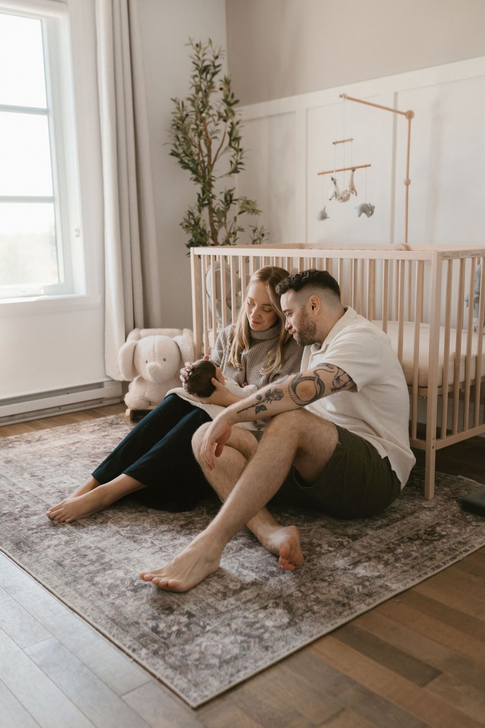 Family sits together on a nursery floor beside a white crib with decorative mobile.
