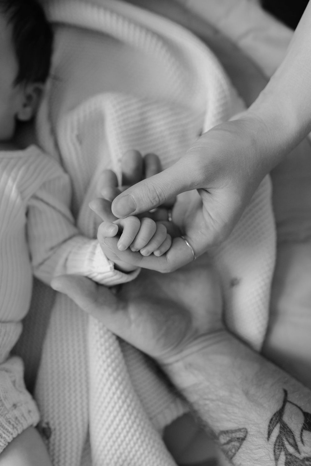 Black and white close-up of tiny baby hand being held by parent with tattoo.