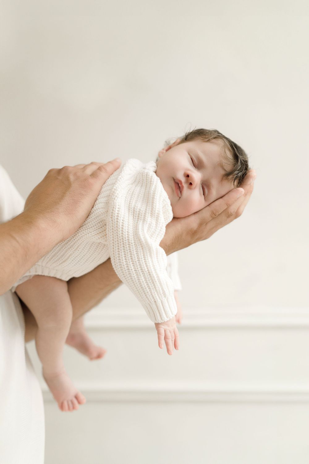 Sleeping baby in white knit outfit being carefully held against light background.