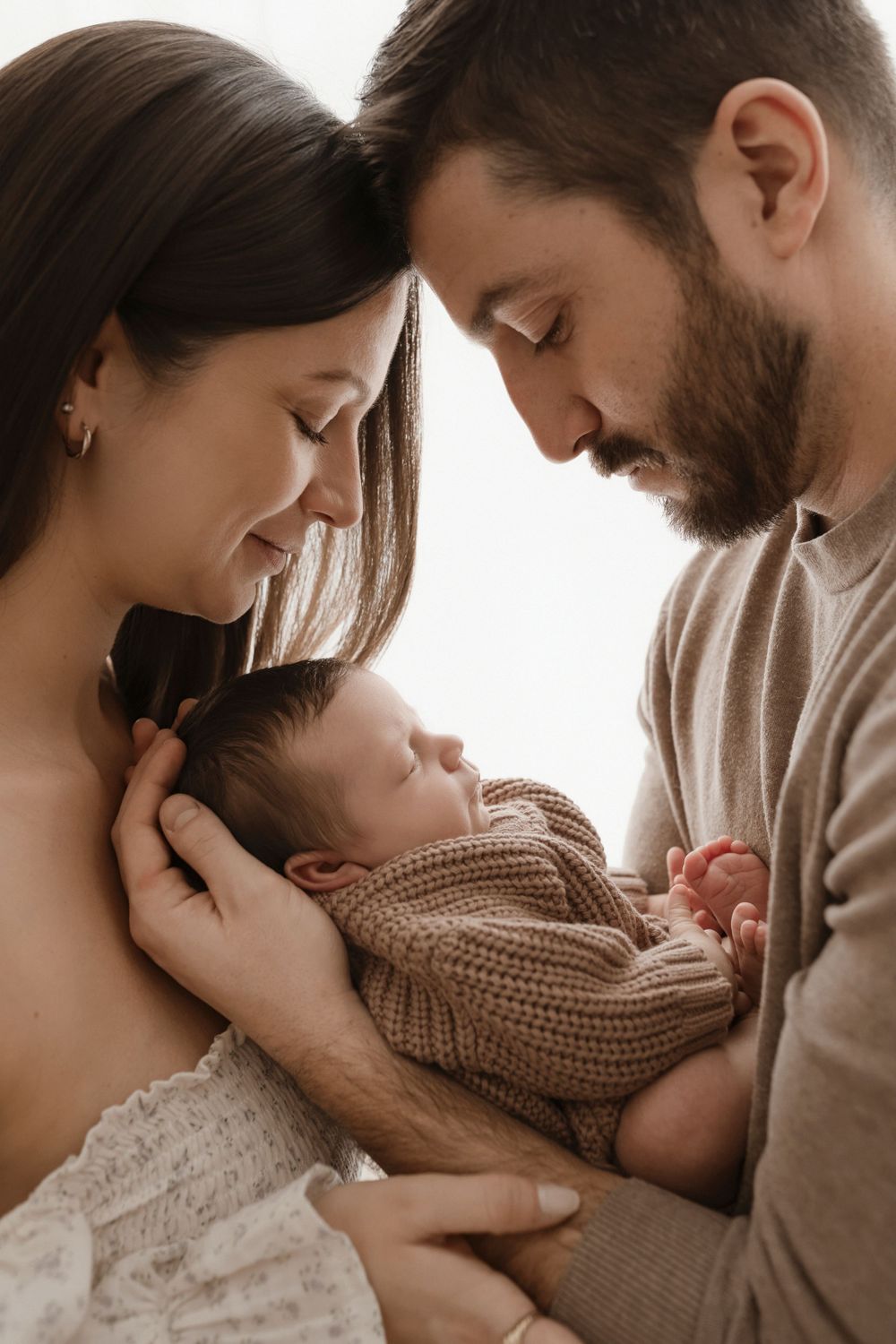 Intimate family portrait with sleeping baby in brown knit sweater against white background.