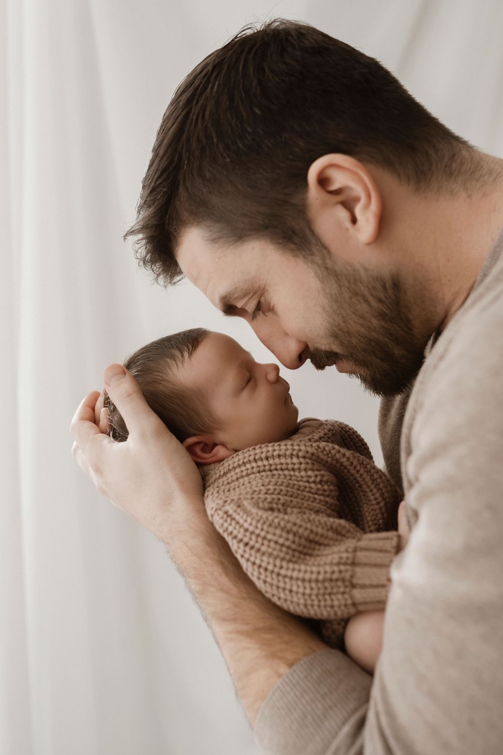 Tender moment with sleeping newborn wrapped in knit blanket against white backdrop.