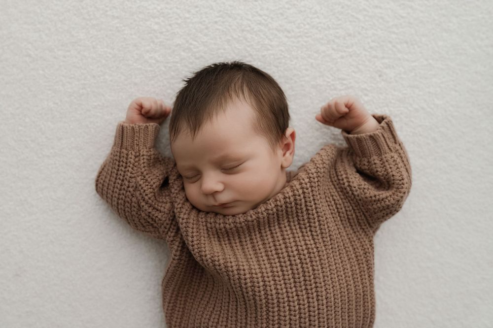 Peaceful sleeping newborn stretches arms while wearing brown knit sweater against white background.
