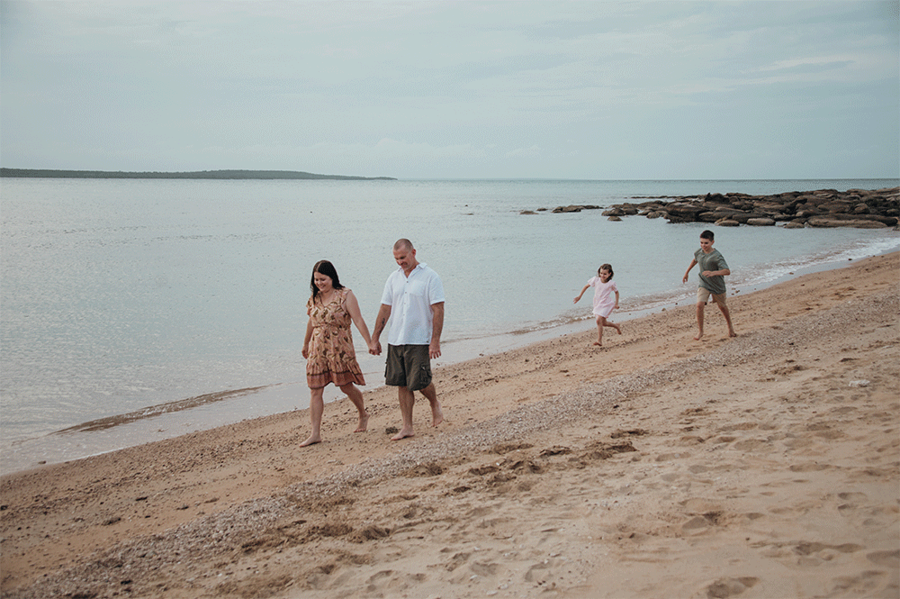 A family walks together along a peaceful sandy beach with rocky coastline and calm waters on a cloudy day.