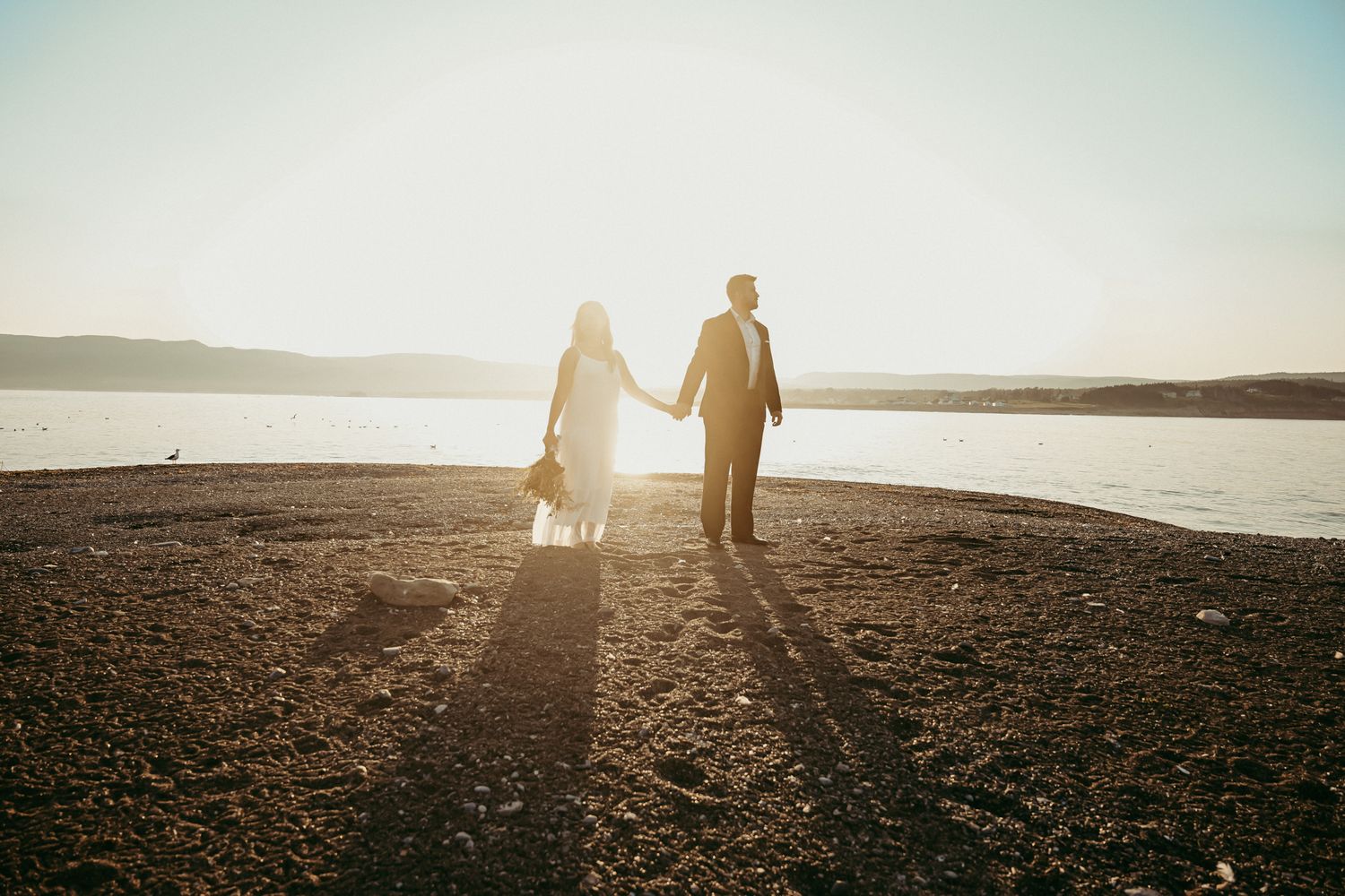 Backlit couple creates long shadows while standing together at sunset by a lake.