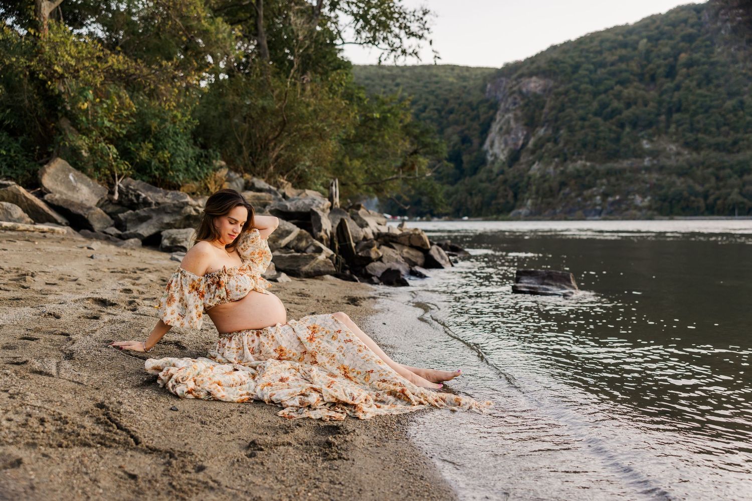 Pregnant woman in hat sitting among wildflowers in rural field at sunset.