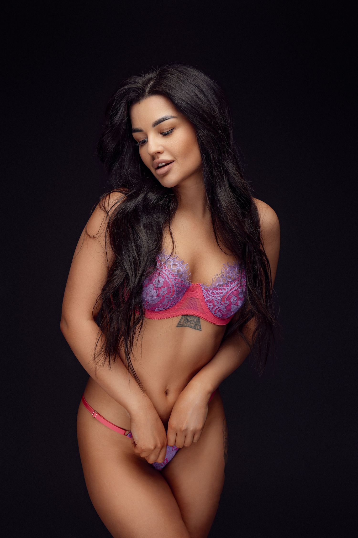 Model in pink and purple lace lingerie set poses against dark background.