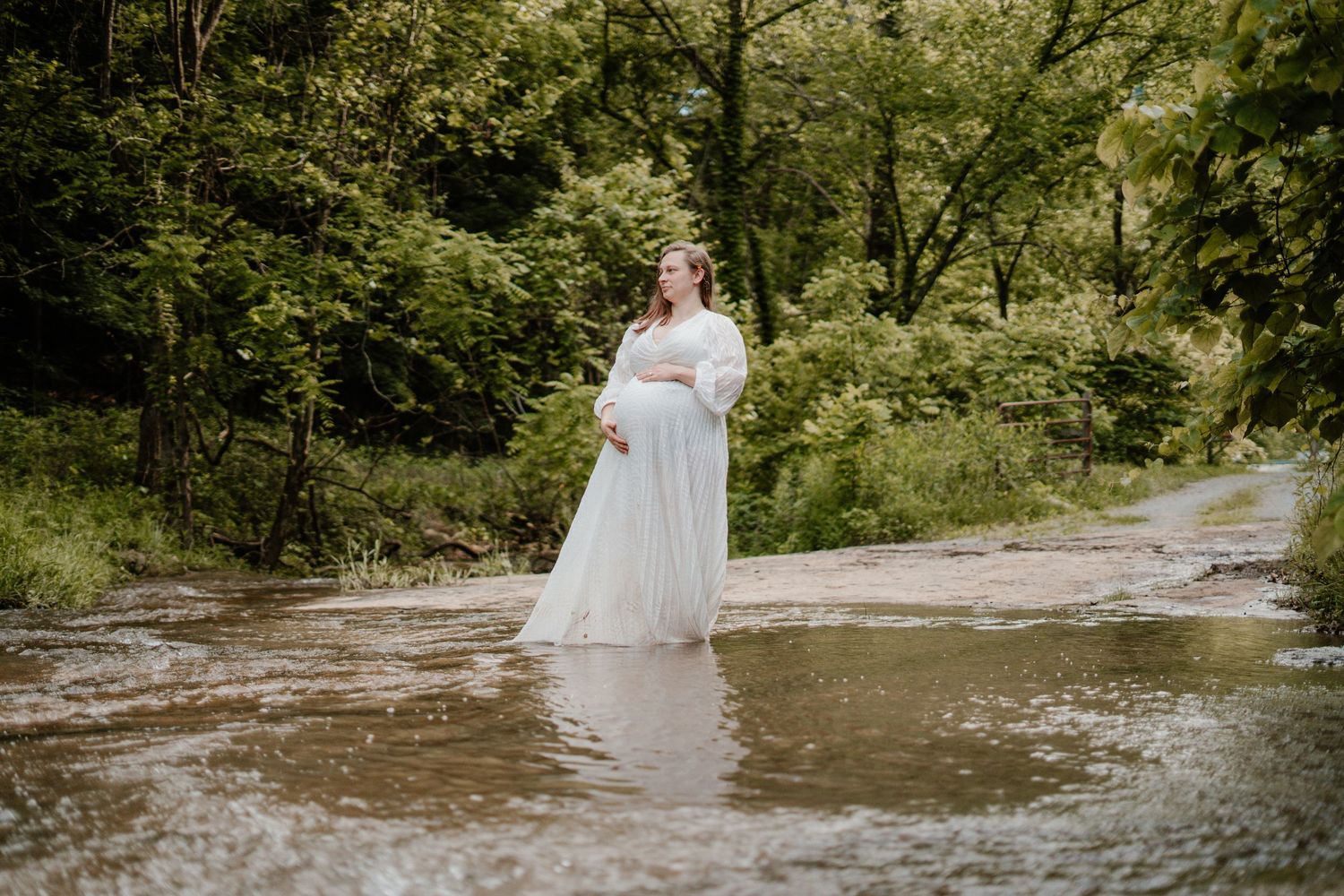 Pregnant woman in flowing white gown standing in a shallow woodland creek surrounded by greenery.
