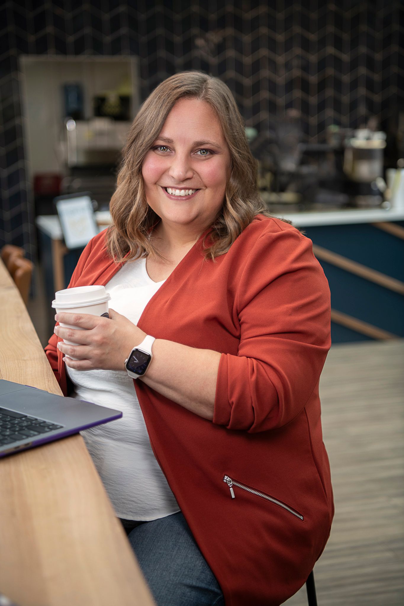 Person in orangejacket holding coffee cup while smiling warmly in modern cafe setting.