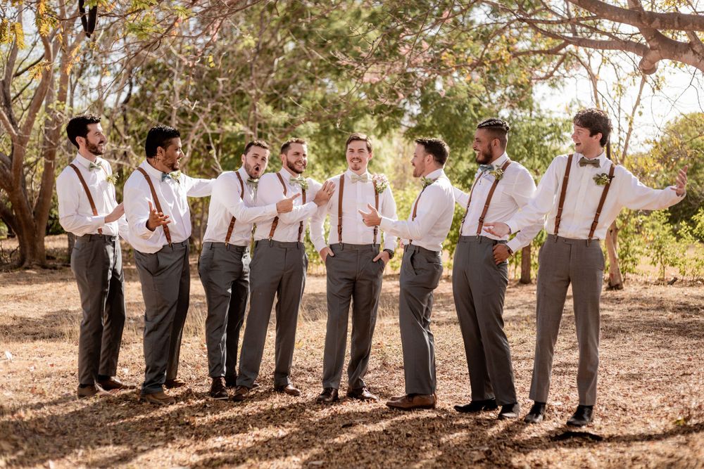 Groomsmen in white shirts and brown suspenders with gray trousers laughing together outdoors at a wedding. Guanacaste, Costa Rica