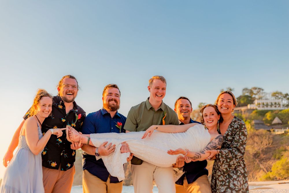 Happy wedding guests playfully lift bride horizontally in the air at outdoor beach celebration. Guanacaste, Costa Rica
