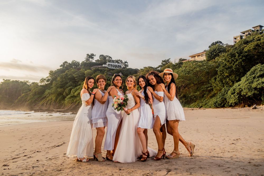 Bride with bouquet and bridesmaids in white dresses smiling together on a scenic beach at sunset. Guanacaste, Costa Rica