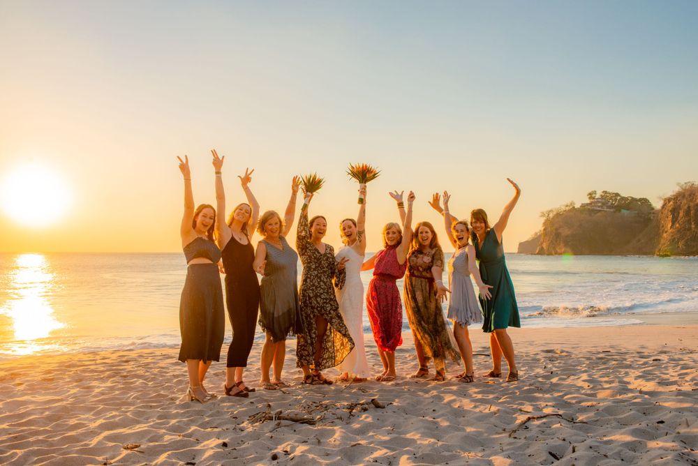 Group of women celebrating at sunset beach wedding, jumping joyfully with bouquet on sandy shore. Guanacaste, Costa Rica