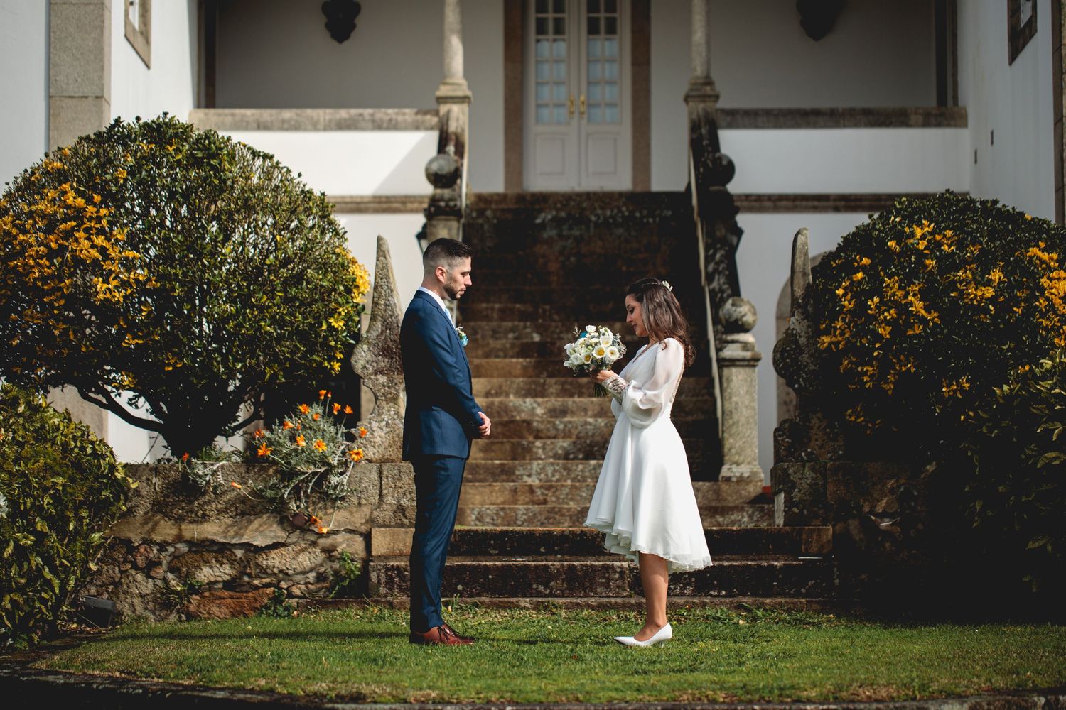 A couple shares a romantic moment on stone steps framed by yellow flowers and manicured shrubs outside a white building.
