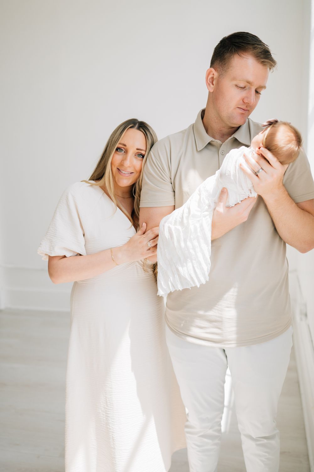 Parents in neutral colors cradle their newborn baby in minimalist white room setting.