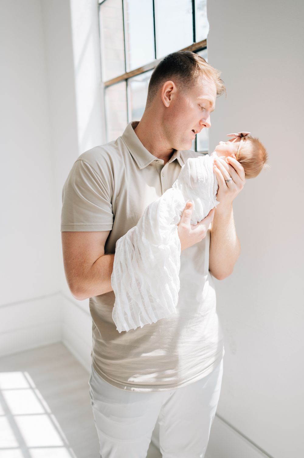 A person in beige holds a newborn baby while standing by a bright window.