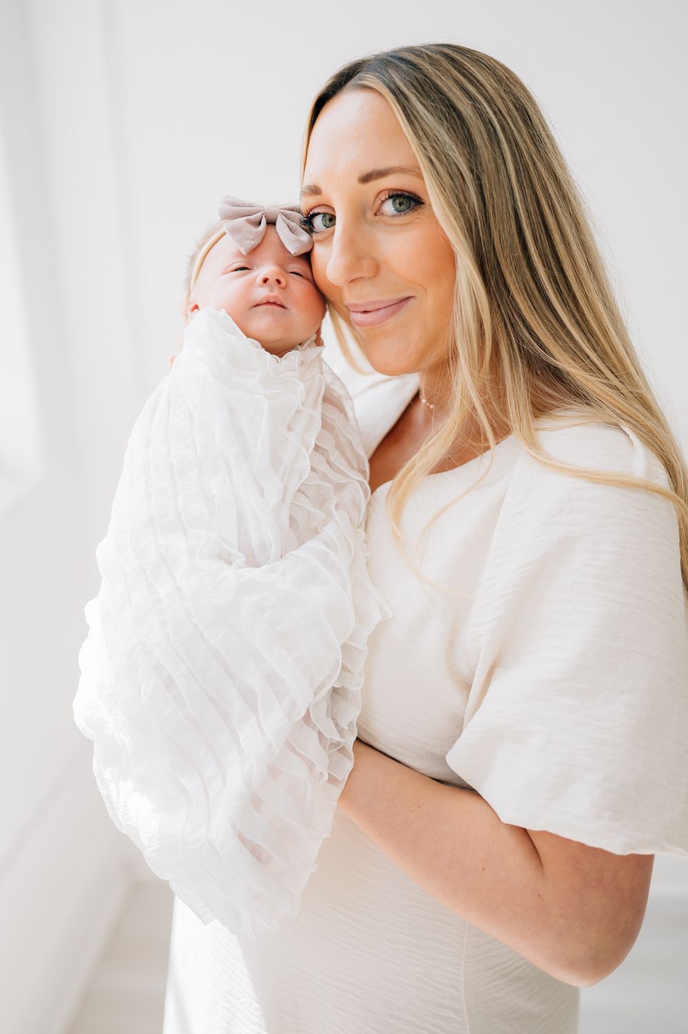 Mother in cream dress holds baby in white outfit against bright white wall.
