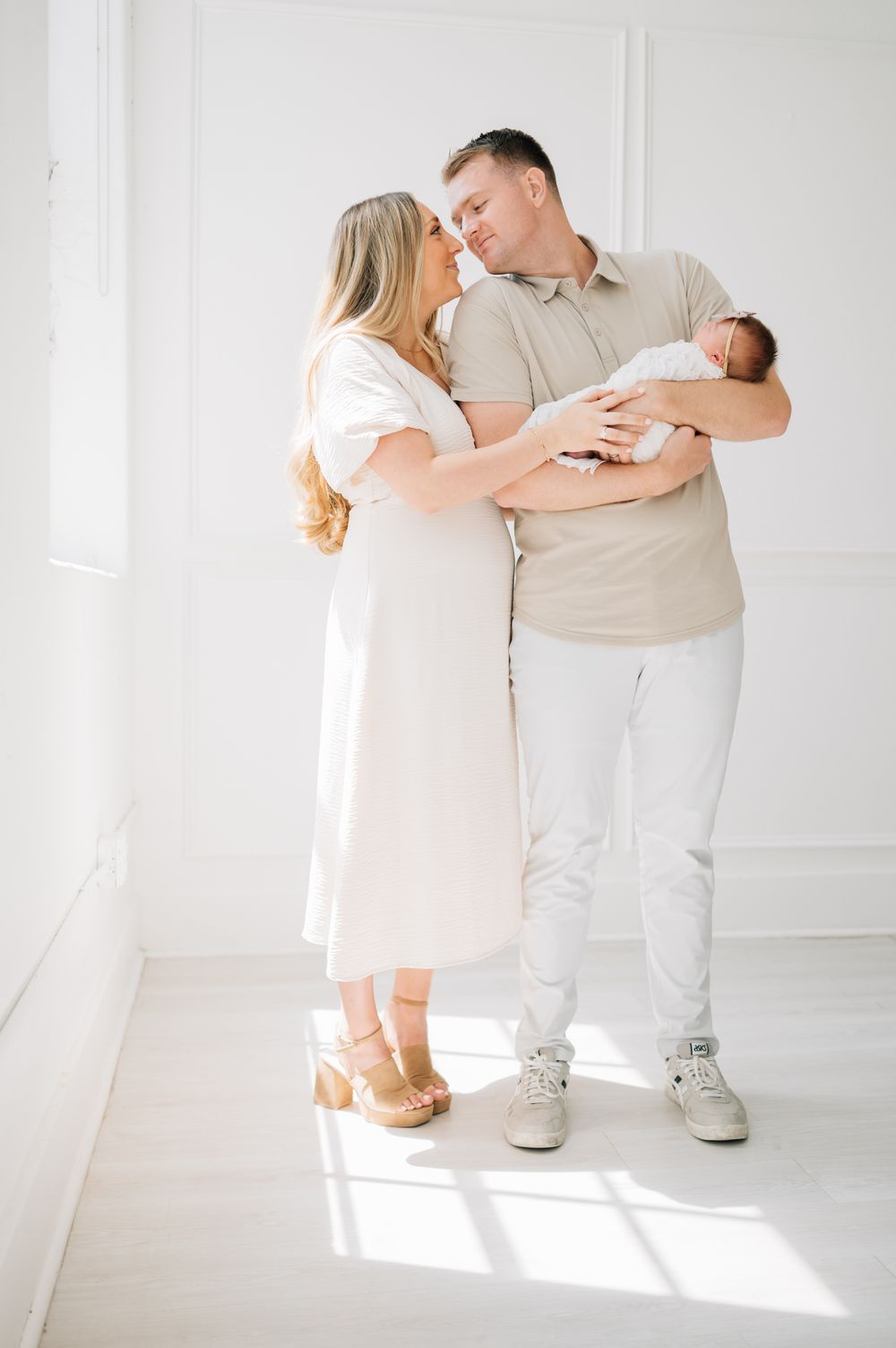 A couple in white and beige outfits stands together with their newborn in a bright room.
