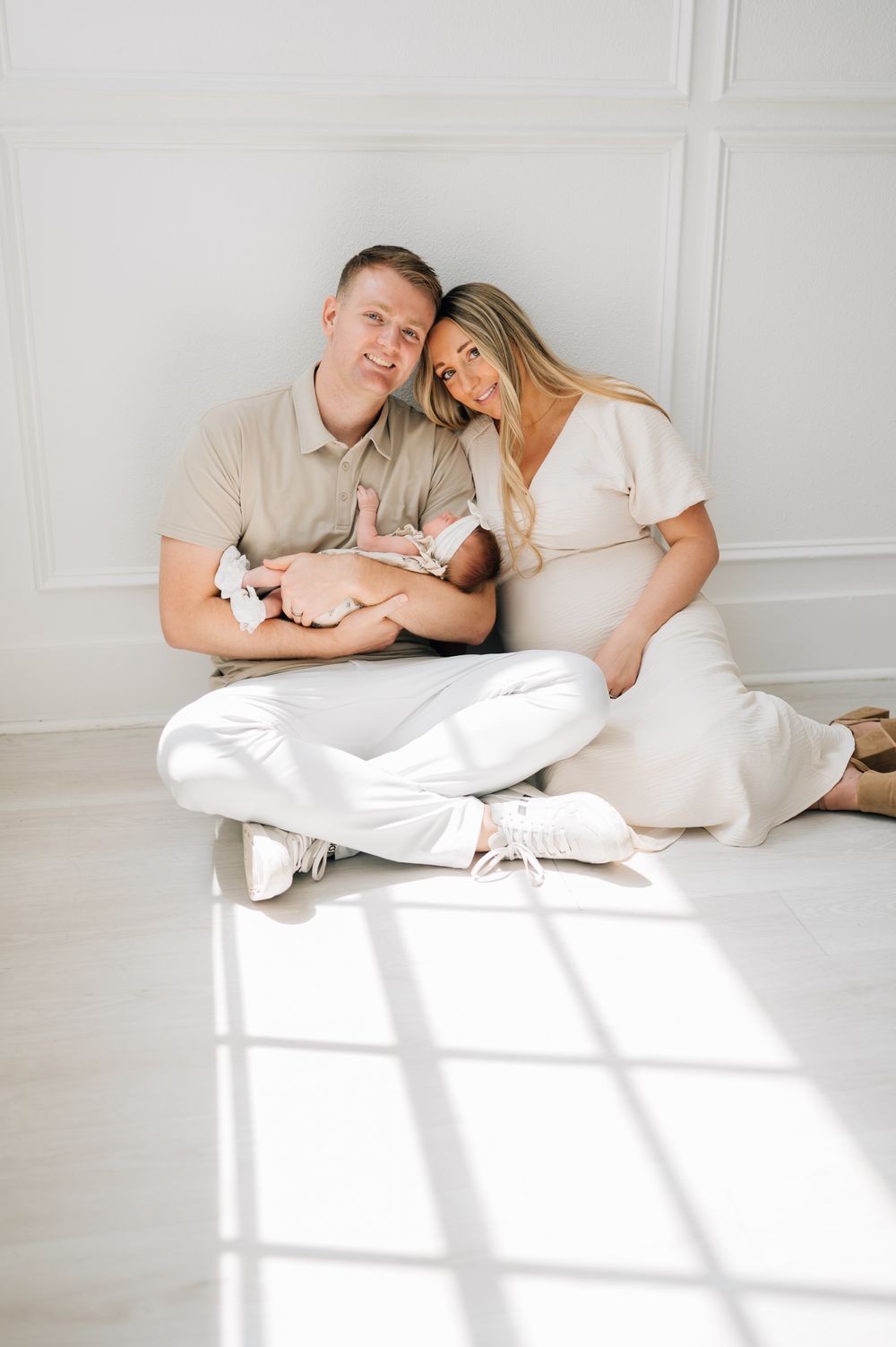 A family sits together on a white floor holding their newborn baby in a minimalist setting.