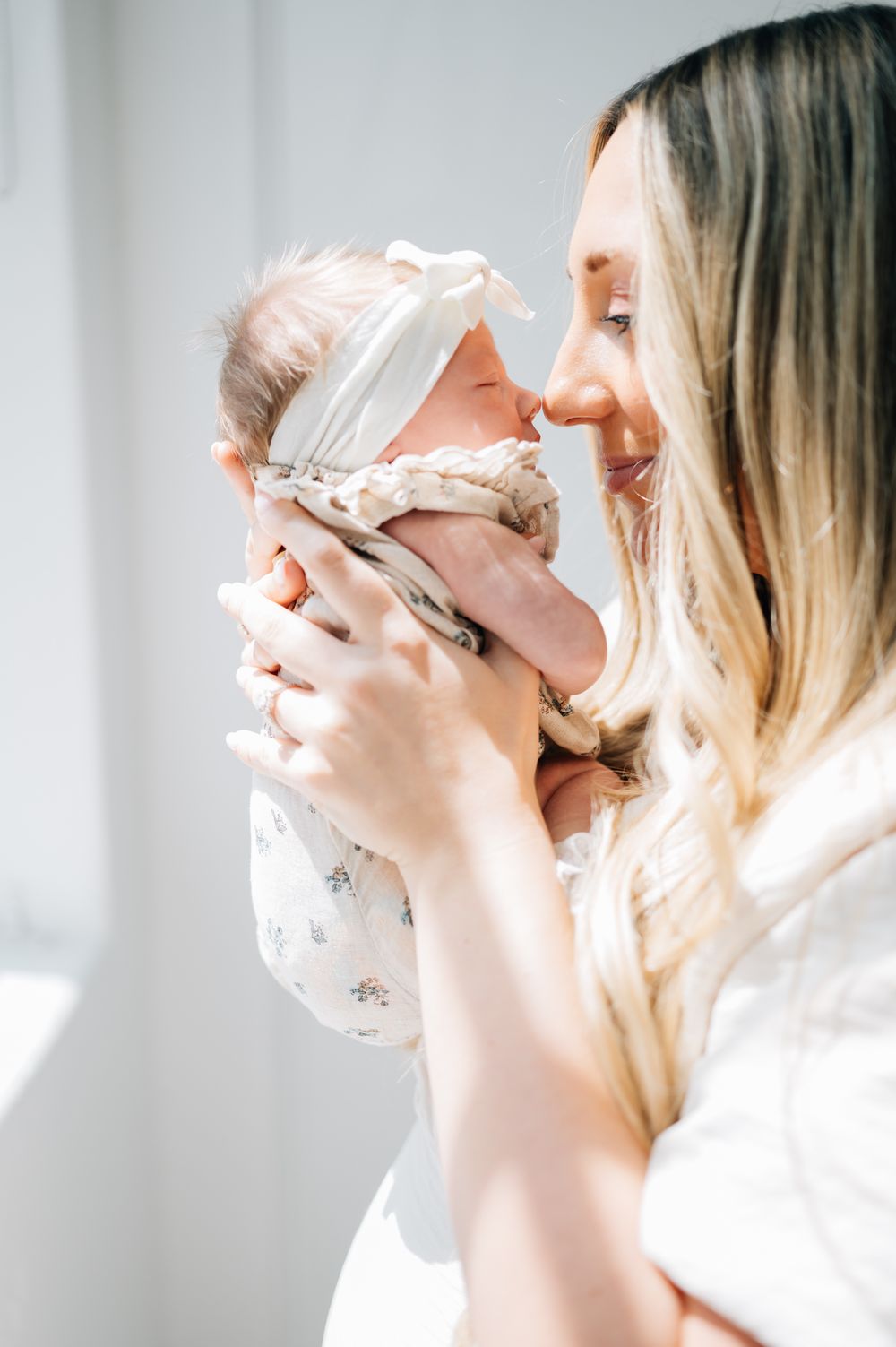 Mother in white dress holds and gazes lovingly at newborn baby in white headband.