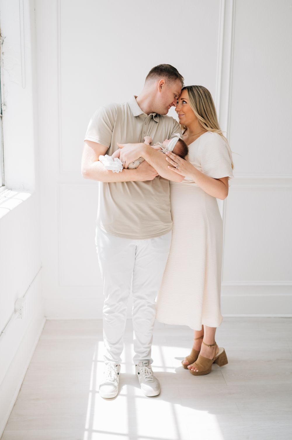 Couple in light colored clothing share intimate moment while holding their newborn in bright room.