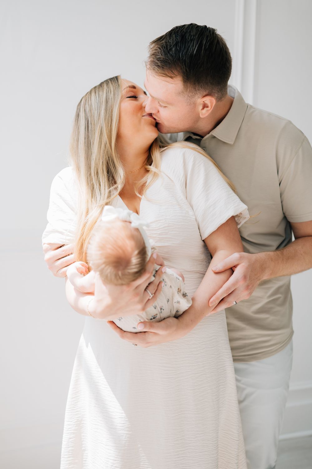 A couple shares an intimate moment while holding their newborn baby in a white room.
