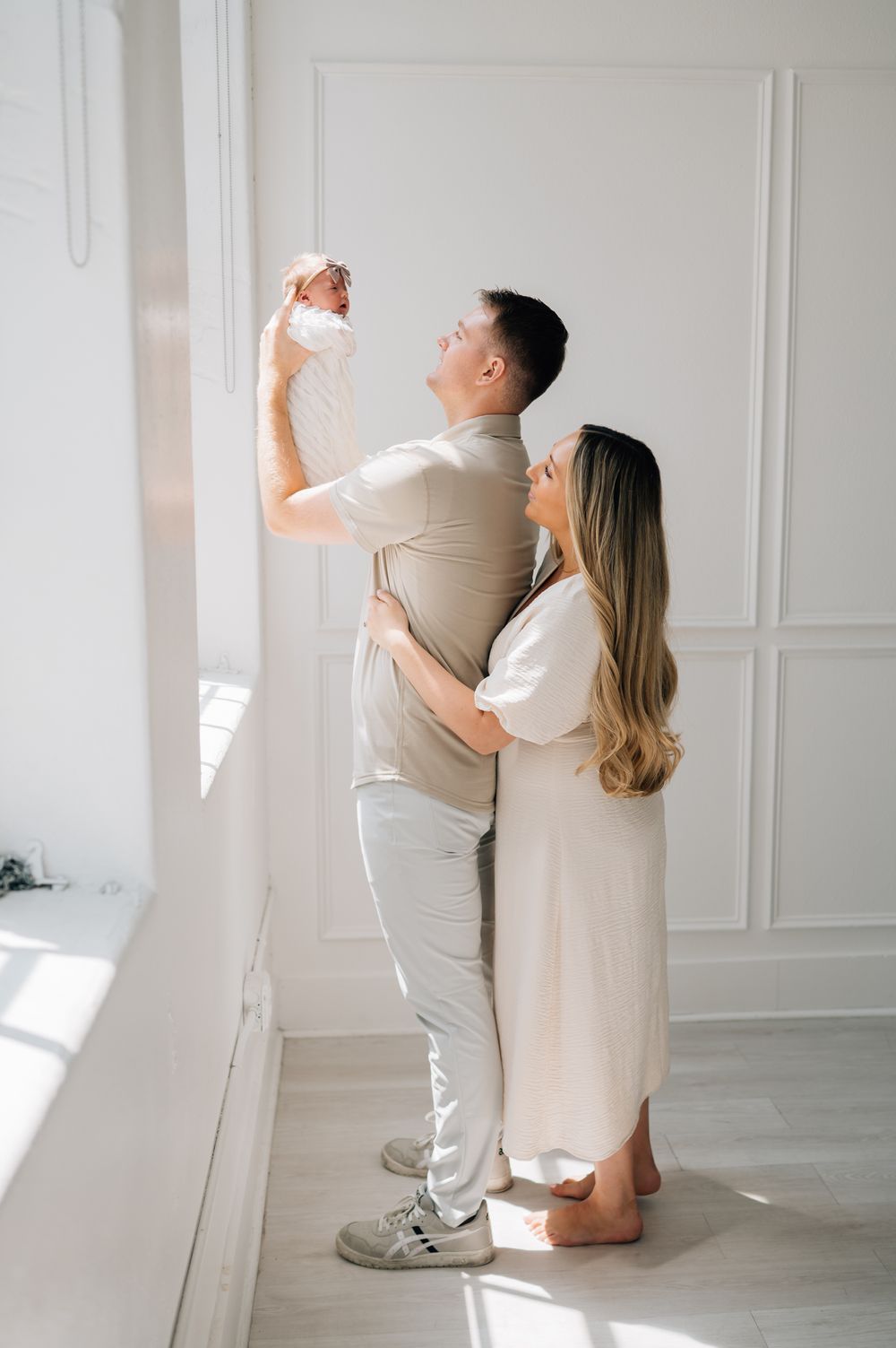 Parents in light clothing lift baby up towards sunlight in minimalist white room.