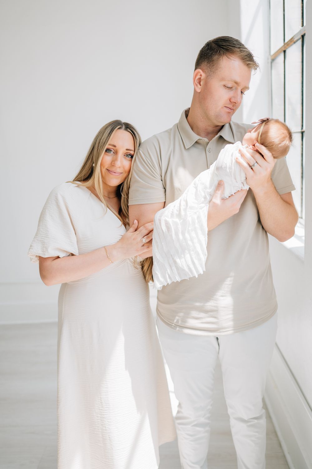 A couple stands by a window cradling their newborn baby in an all-white room.