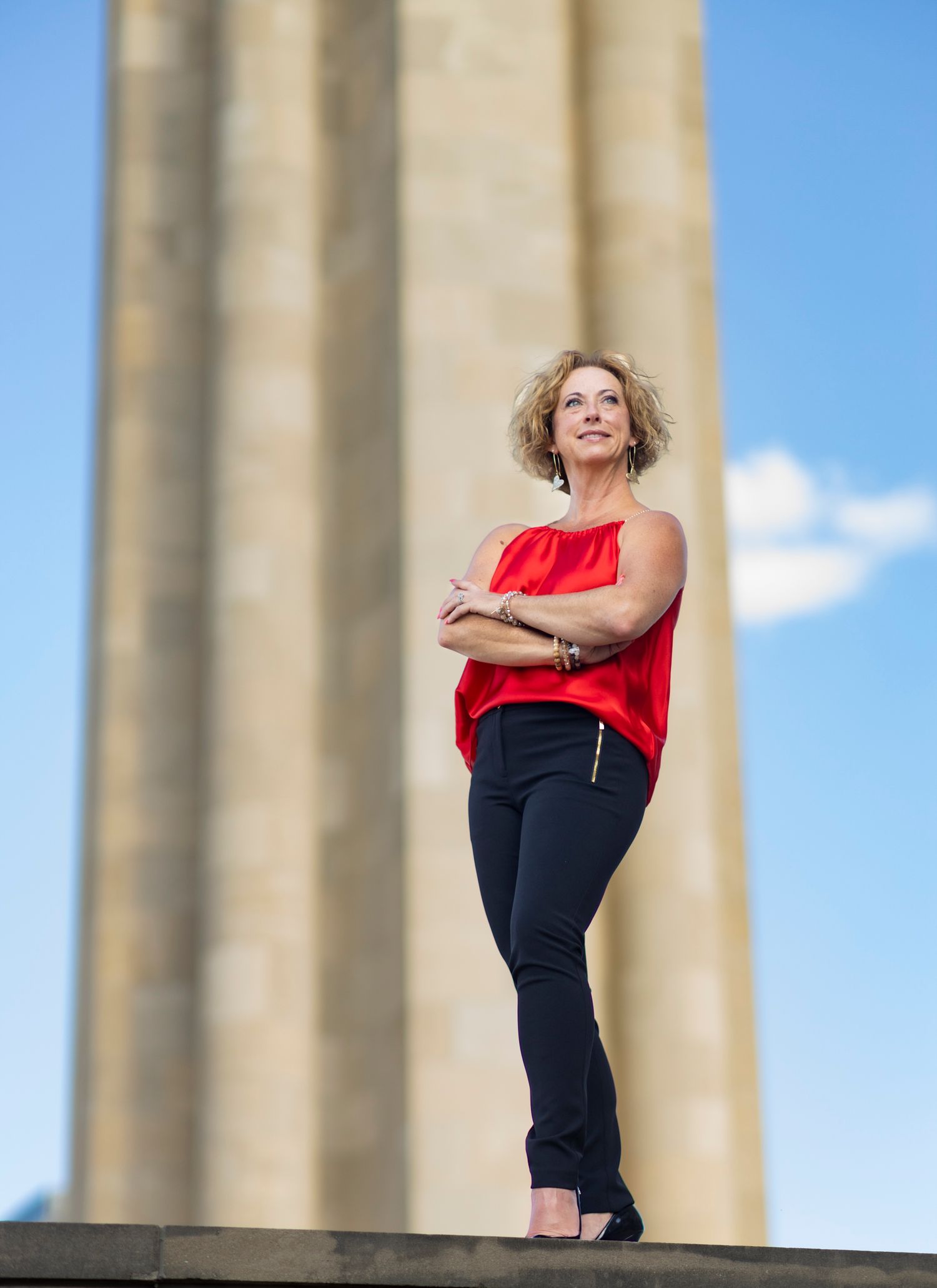 Woman in red top and dark pants poses confidently against classical stone columns.