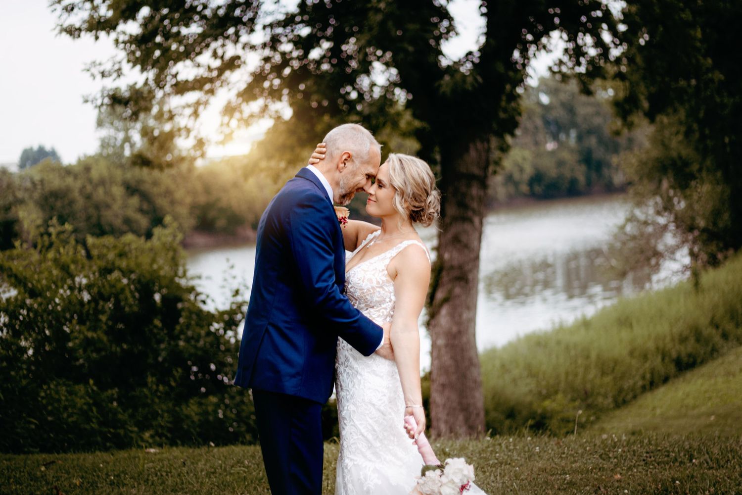 Bride and groom share a romantic moment by a sunlit lake, surrounded by lush greenery at their outdoor wedding.