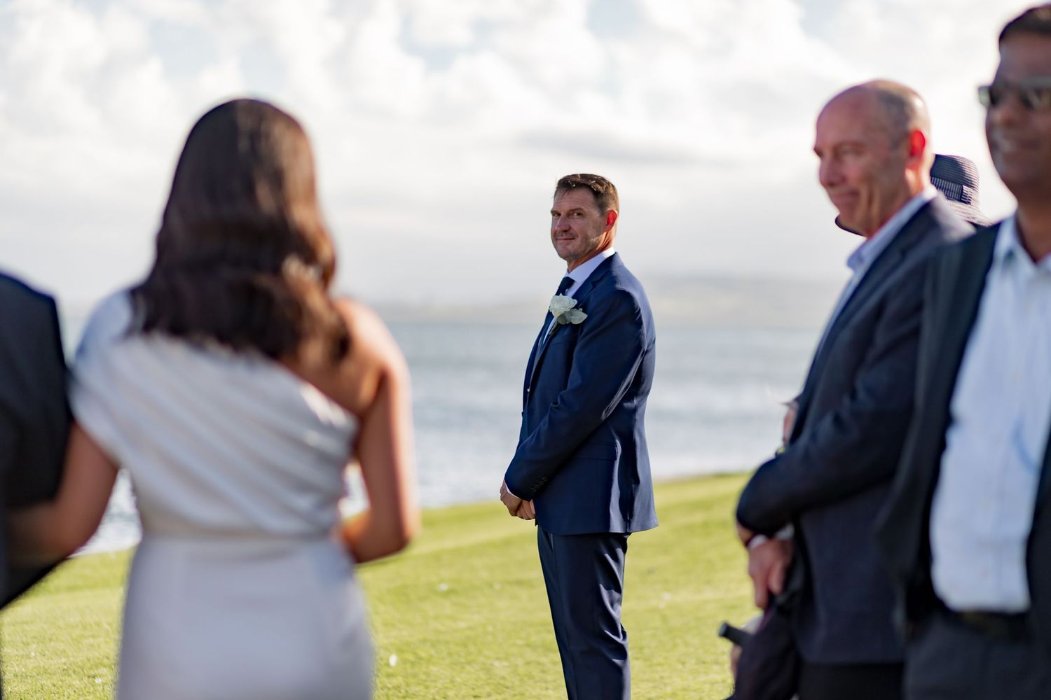 Wedding ceremony participants standing on grass with ocean view in background.