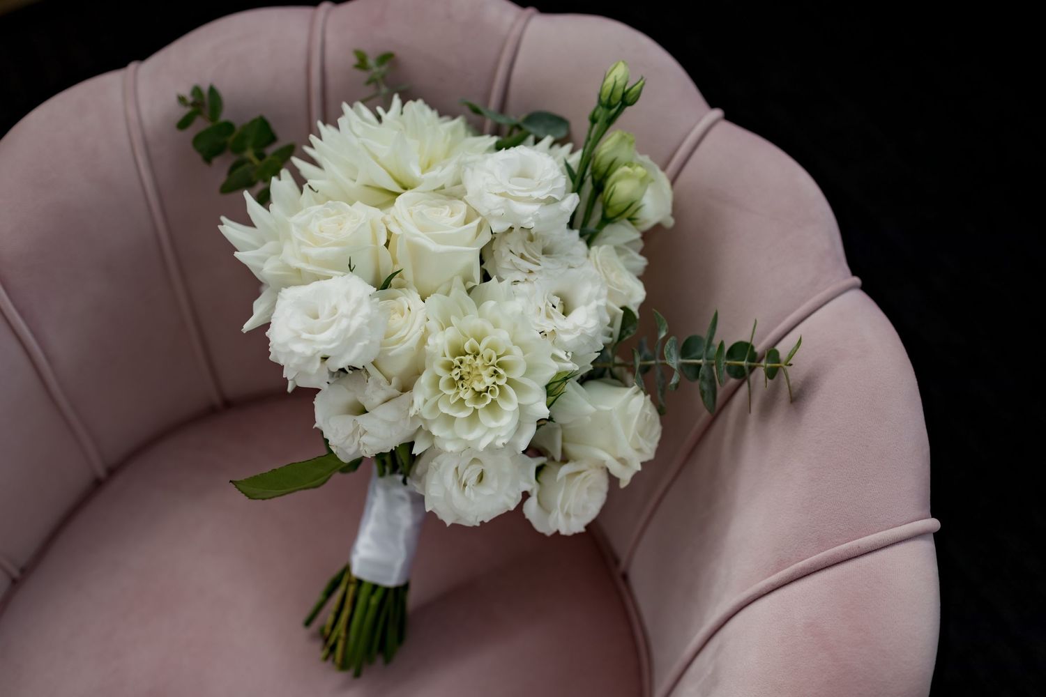 Elegant white floral wedding bouquet with roses and greenery placed on pink velvet chair.