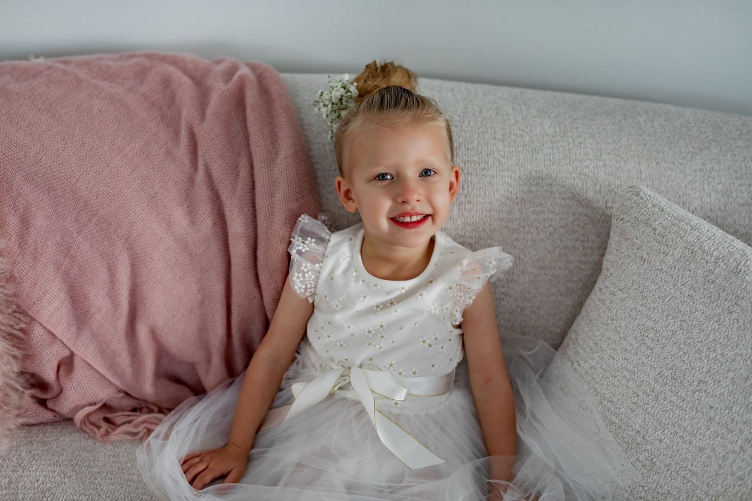 Young child in white dress sitting on grey couch with pink throw blanket.