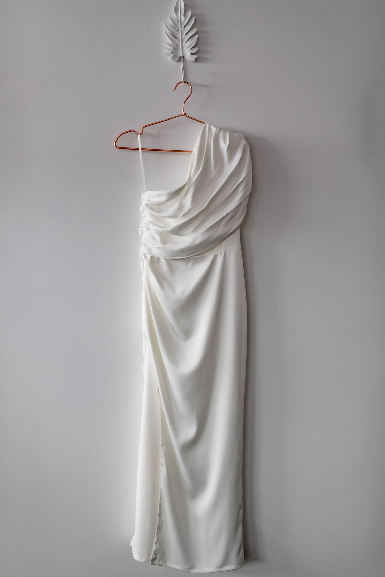 Elegant white one-shoulder wedding dress hanging on copper hanger with feather wall decoration.