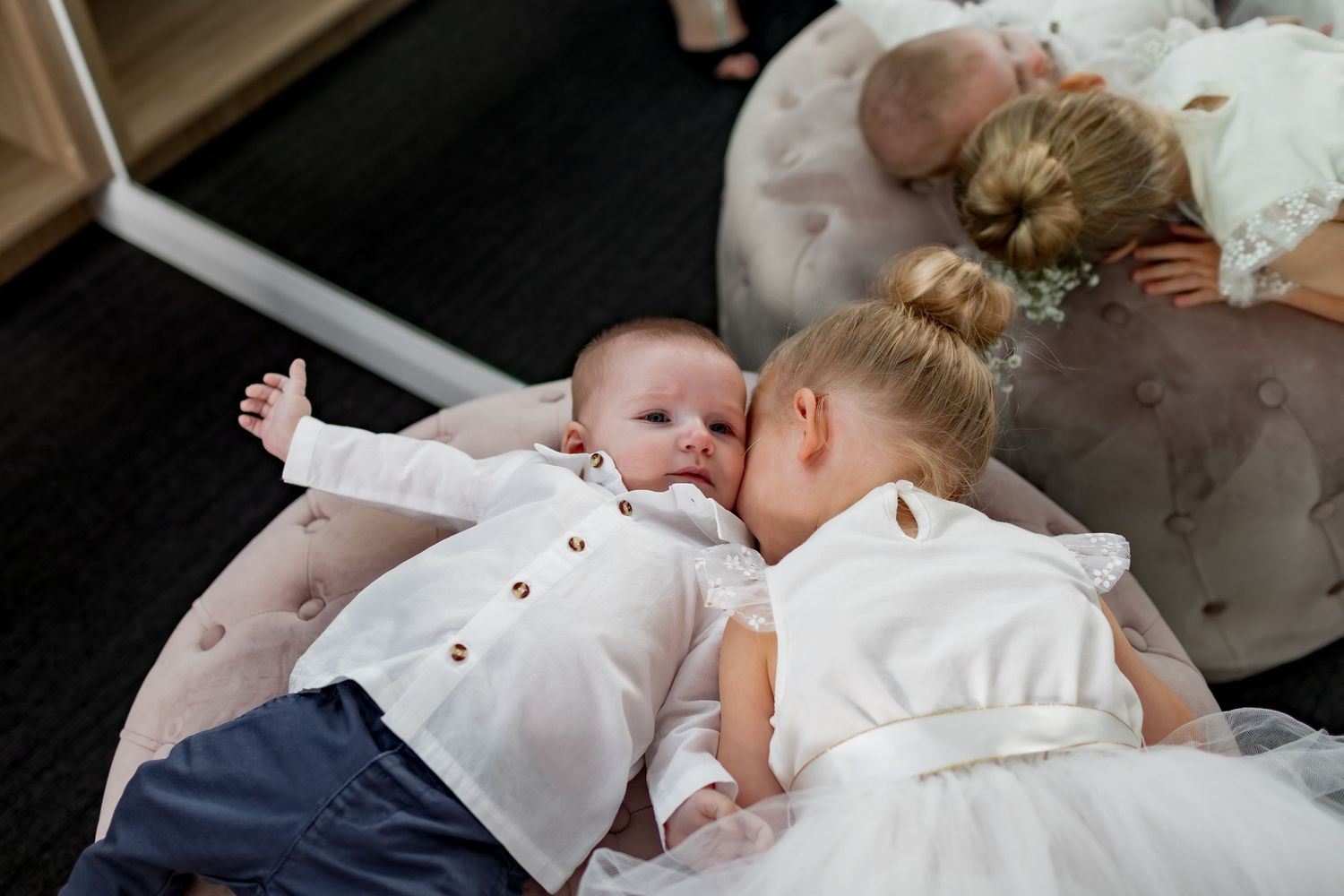 Two children in formal white attire laying together on dark floor.