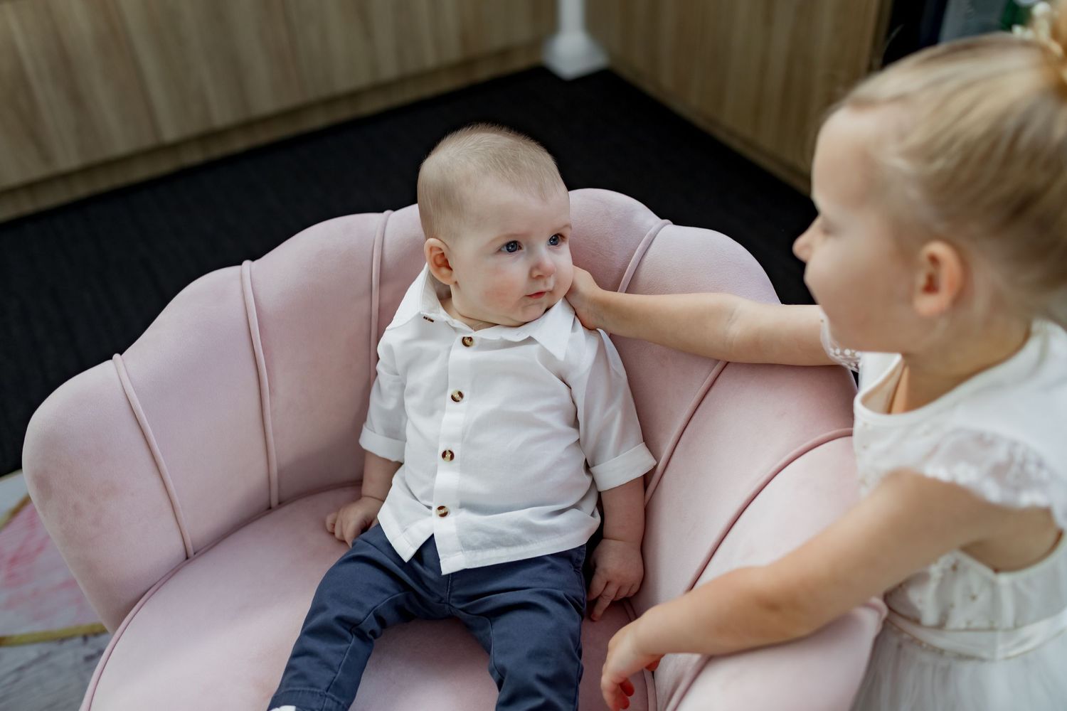 Two children interacting on pink velvet chair wearing formal white outfits.